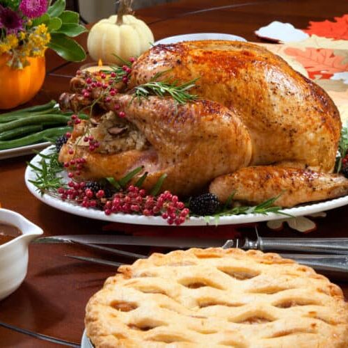 Roasted pepper turkey for Thanksgiving, garnished with pink pepper, blackberry, and fresh rosemary twigs on a dinner table decorated with mini pumpkins, beans, carrots, baked potato, pie, cranberry relish, gravy, flowers, candles, and flutes of champagne."n