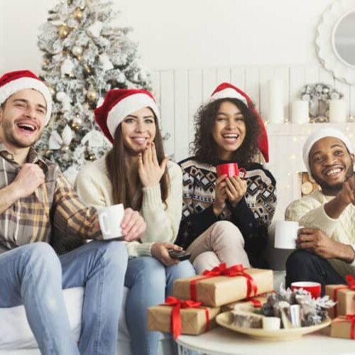 Christmas comedy show. Happy laughing friends in Santa hats watching Tv together at home, copy space